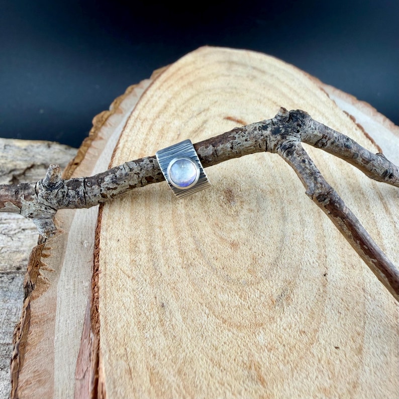 K&ouml;nnte beinhalten: Ein Silberring mit strukturiertem Band und einem runden, irisierenden Stein. Der Ring wird auf einem kleinen, verwitterten Ast vor einem Holzgrund pr&auml;sentiert, der seine handwerklichen Details und sein einzigartiges Design zeigt.