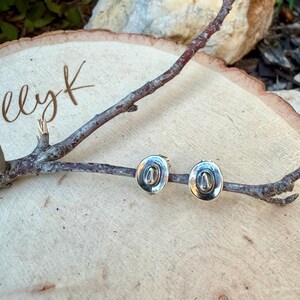 May include: A pair of silver-coloured cowboy hat stud earrings. The earrings are displayed on a wooden branch, set against a light-coloured wooden background. The earrings have a detailed design.