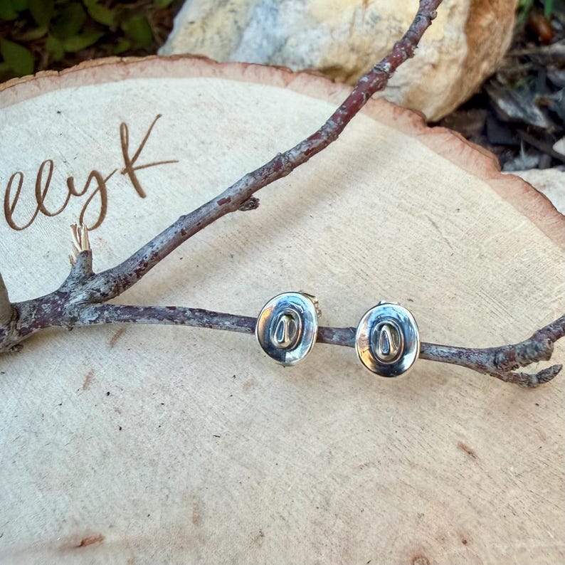 May include: A pair of silver-coloured cowboy hat stud earrings. The earrings are displayed on a wooden branch, set against a light-coloured wooden background. The earrings have a detailed design.