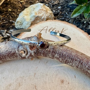 Puede incluir: Brazalete de plata con textura martillada y una piedra preciosa de color rojo oscuro engastada en un bisel dorado. El brazalete se exhibe sobre una superficie de madera con la palabra "Allink" escrita.