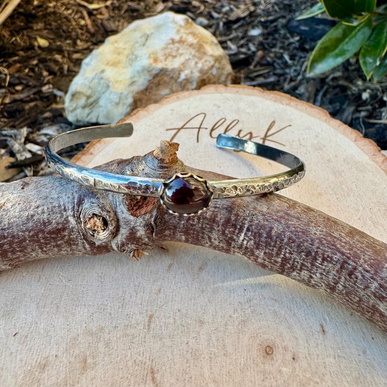 Puede incluir: Brazalete de plata con textura martillada y una piedra preciosa de color rojo oscuro engastada en un bisel dorado. El brazalete se exhibe sobre una superficie de madera con la palabra "Allink" escrita.