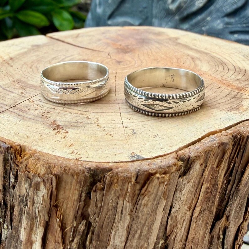 May include: Two silver rings with engraved geometric patterns and beaded edges. The rings feature a polished metallic finish and intricate stamped detailing along the outer bands, showcasing a consistent design across both circular pieces.