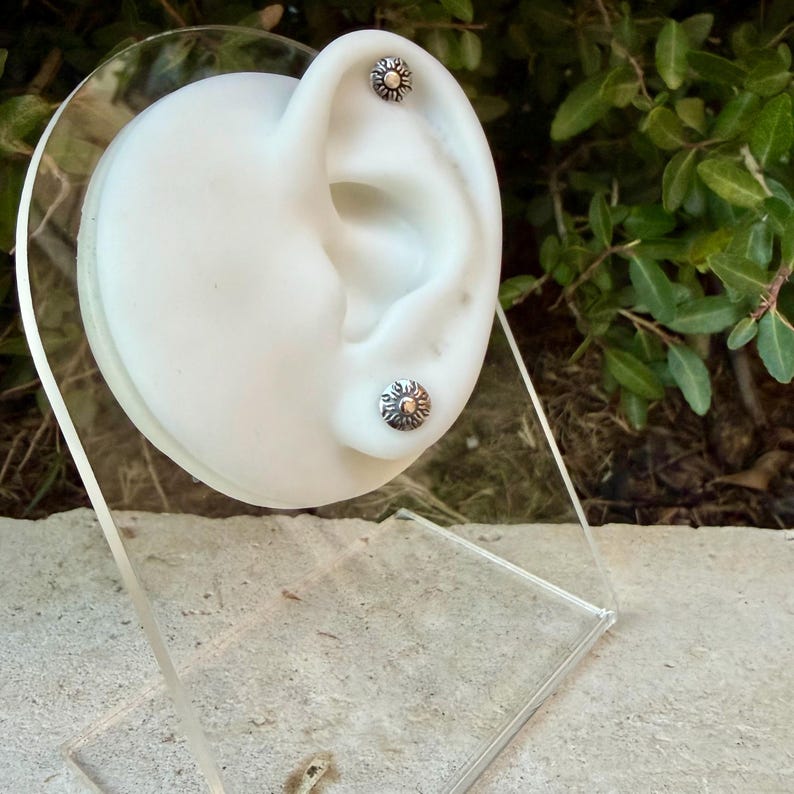 May include: Two silver stud earrings shaped like small suns with radiating rays and a central circular detail. The earrings are displayed on a white ear-shaped model, with one stud positioned in the upper cartilage and one in the earlobe.