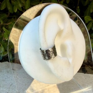 Puede incluir: Un pendiente tipo ear cuff de tono plateado con textura martillada y un patr&oacute;n de cruz geom&eacute;trica central. La banda de metal tiene un dise&ntilde;o ancho y abierto que se ajusta al borde exterior de un modelo de oreja blanco de exhibici&oacute;n.