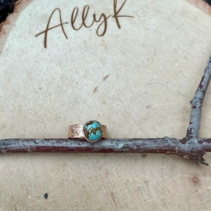 May include: A gold-colored ring with a turquoise stone. The ring is set on a wooden surface with the name "Allyk" written in cursive. The ring's band has a textured design. The turquoise stone has brown and blue patterns.