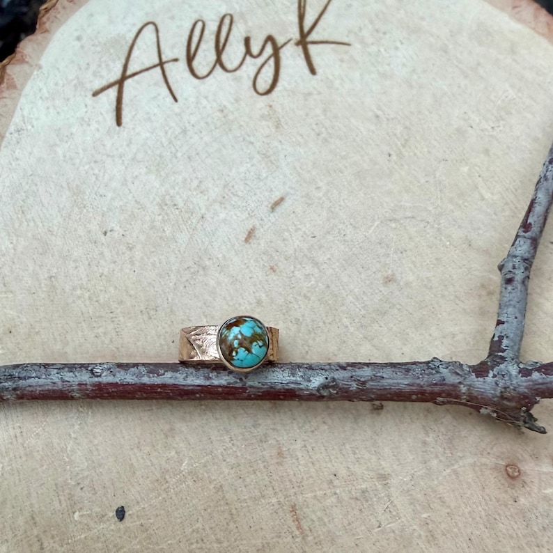 May include: A gold-colored ring with a turquoise stone. The ring is set on a wooden surface with the name "Allyk" written in cursive. The ring's band has a textured design. The turquoise stone has brown and blue patterns.