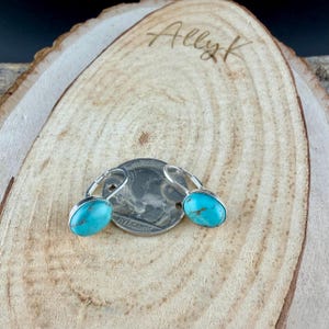 May include: A pair of turquoise leverback earrings with silver-colored metal settings. The earrings are displayed on a wooden surface with the handwritten text "Ally K". A US nickel coin is also present.