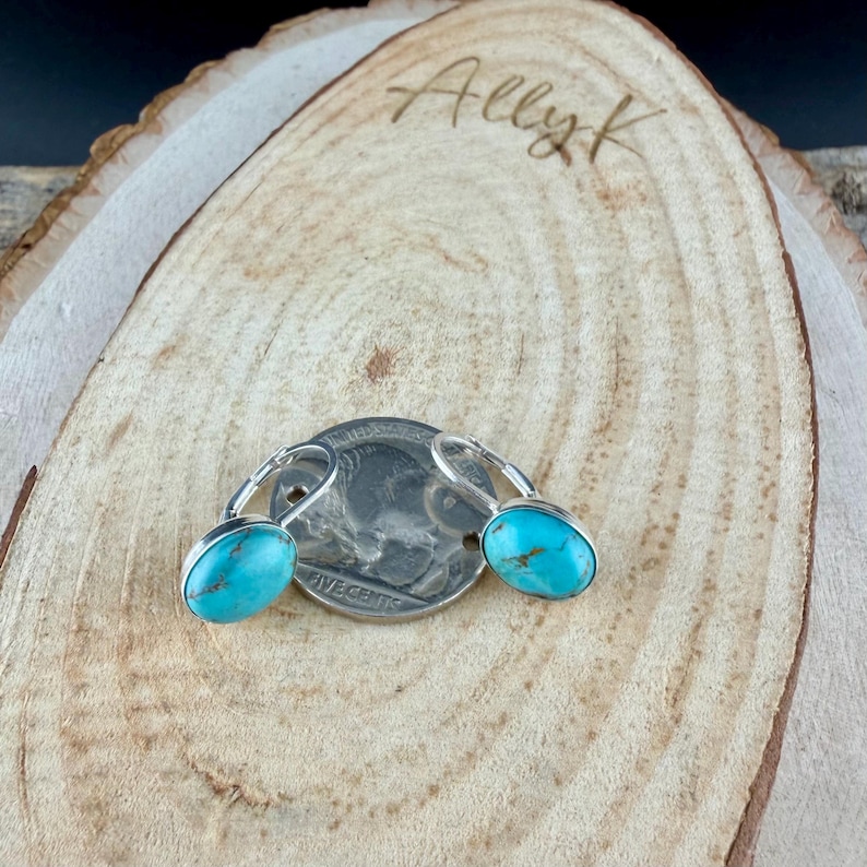 May include: A pair of turquoise leverback earrings with silver-colored metal settings. The earrings are displayed on a wooden surface with the handwritten text "Ally K". A US nickel coin is also present.