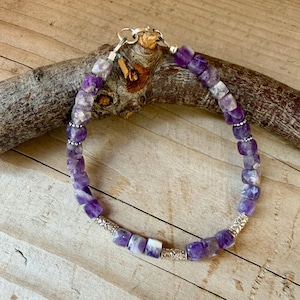 Puede incluir: Una pulsera con cuentas c&uacute;bicas de amatista morada facetada dispuestas en un patr&oacute;n circular. El dise&ntilde;o incluye peque&ntilde;as cuentas espaciadoras de metal plateado con grabados florales y un cierre de mosquet&oacute;n de metal plateado.