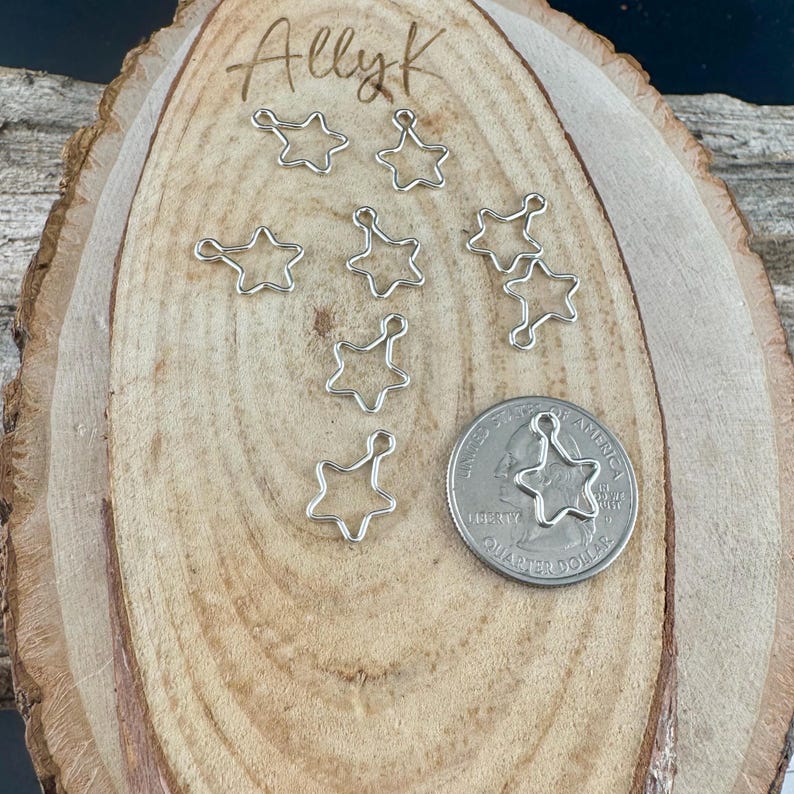 May include: A collection of small, silver-colored star charms with a loop at the top, scattered on a wooden surface. A US quarter coin is included for size comparison. The word "Allyk" is written in cursive at the top.