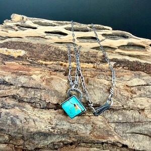 May include: A silver necklace with a square turquoise pendant. The pendant is set in a silver frame and hangs from a delicate chain. The necklace is displayed on a textured wooden surface.