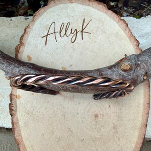 May include: A copper-colored, twisted metal cuff bracelet. The bracelet is displayed on a wooden branch and a light-colored wooden slice. The text "AllyK" is written on the wooden slice.