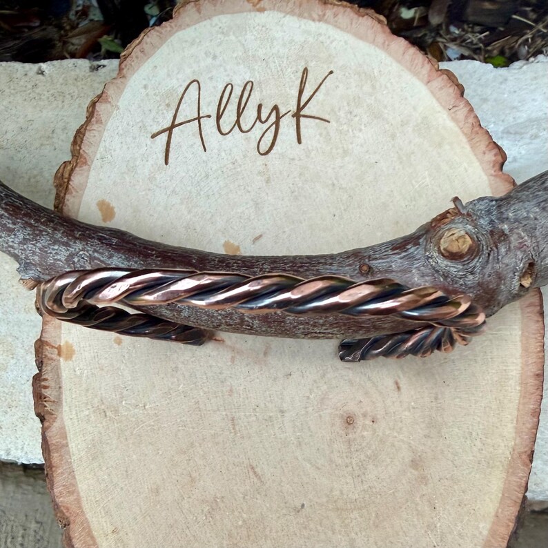 May include: A copper-colored, twisted metal cuff bracelet. The bracelet is displayed on a wooden branch and a light-colored wooden slice. The text "AllyK" is written on the wooden slice.