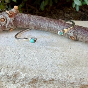 May include: A silver cuff bracelet featuring two small, oval-shaped turquoise stones set at each open end. The metal band has a smooth, polished finish, and the turquoise stones display a vibrant blue-green colour with dark matrix veining.