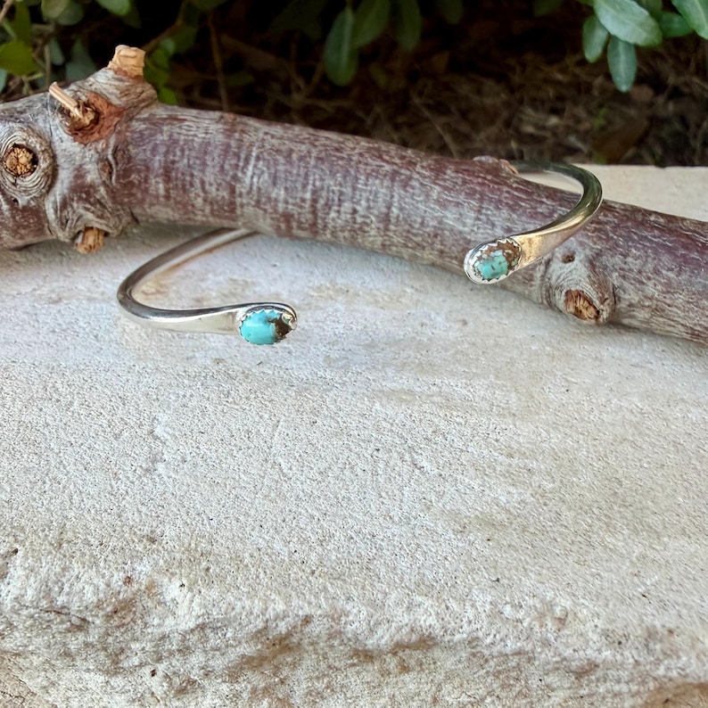 May include: A silver cuff bracelet featuring two small, oval-shaped turquoise stones set at each open end. The metal band has a smooth, polished finish, and the turquoise stones display a vibrant blue-green colour with dark matrix veining.
