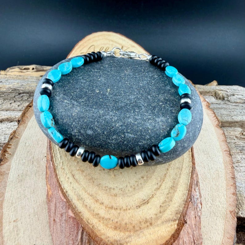 May include: A turquoise and black beaded bracelet with a silver clasp. The bracelet features oval turquoise beads, interspersed with black and silver beads. The bracelet is displayed on a gray stone.