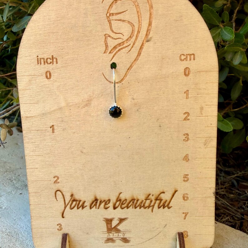 May include: A silver-toned earring with a small, round, black stone, displayed on a light-colored wooden card featuring an etched ear outline, a ruler with inch and centimeter markings, and the text "You are beautiful" with a K logo.