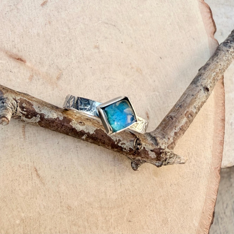 May include: Two silver rings with textured bands and square, blue-green center stones. The rings are displayed on a small, brown branch against a light brown wooden background. The rings are likely jewelry.