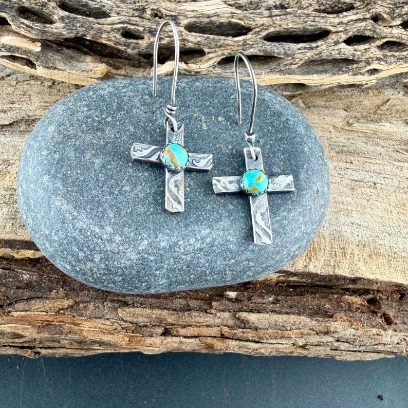 May include: Silver cross earrings with turquoise-colored stone centers. The crosses have etched designs and are displayed on a gray stone. The earrings have long, curved hooks.