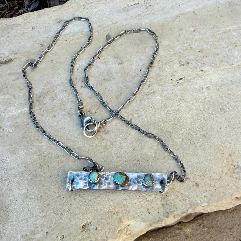 May include: A silver-toned necklace with a hammered bar pendant. The pendant features three turquoise stones. The chain is a linked design with a lobster clasp closure. The necklace is displayed on a stone surface.