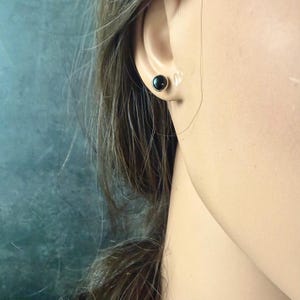 May include: A close-up of a person's ear with a black stud earring. The earring is a small, round, black stone set in a gold-colored metal. The person has long, brown hair.
