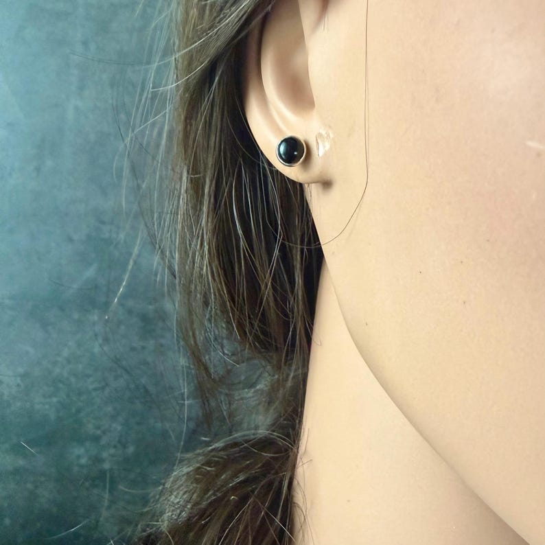May include: A close-up of a person's ear with a black stud earring. The earring is a small, round, black stone set in a gold-colored metal. The person has long, brown hair.