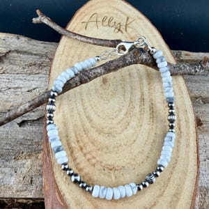 May include: A beaded bracelet featuring white and gray stone beads interspersed with silver-colored metal beads. The bracelet is displayed on a wooden surface with the handwritten text "Allyk". The clasp is silver-colored.
