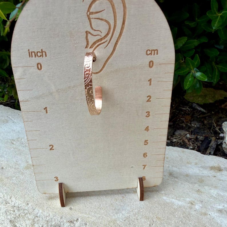 May include: A single rose gold hoop earring with an etched floral design is displayed on a wooden earring display. The display has inch and centimeter measurements. The earring is approximately 1 inch in length.