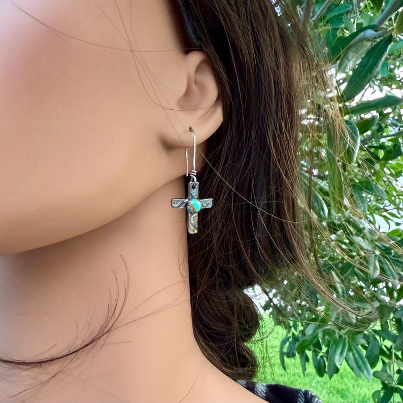 May include: Silver-toned cross earrings with a turquoise-colored stone in the center. The earrings are hanging from a silver hook. The cross has etched details and is worn on the ear.