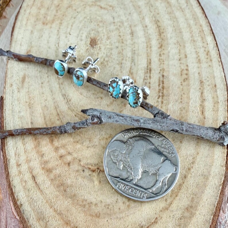 May include: Three pairs of silver stud earrings with turquoise stones are displayed on a small branch. A silver buffalo nickel is also present. The earrings have different designs, including oval and round shapes. The turquoise stones have brown matrix.