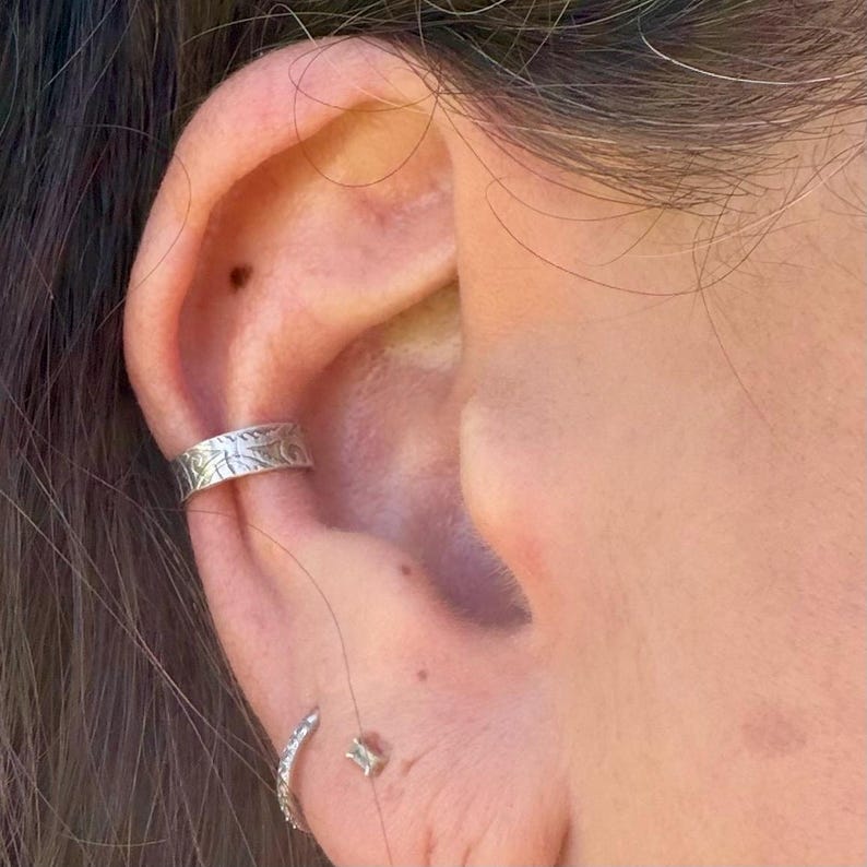May include: Close-up of an ear adorned with silver jewelry. A textured ear cuff with a floral design is clamped on the upper ear. A small, curved earring with clear stones and a small square stud earring are also visible.