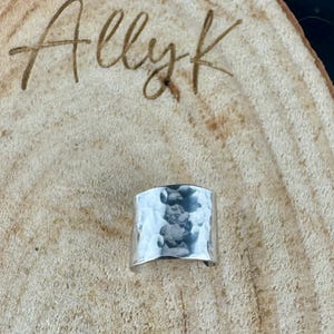 May include: A wide, hammered silver ring. The ring has a textured surface with a slightly curved shape. The ring is displayed on a light brown wooden surface. The text "Allyk" is written in cursive in the background.