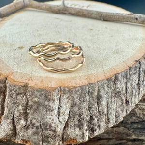 May include: Two gold-colored wavy rings are stacked on a wooden surface. The rings have a smooth, polished finish and a simple, elegant design. The rings are likely made of metal and are suitable for everyday wear.