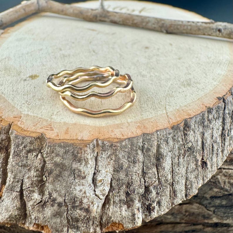 May include: Two gold-colored wavy rings are stacked on a wooden surface. The rings have a smooth, polished finish and a simple, elegant design. The rings are likely made of metal and are suitable for everyday wear.