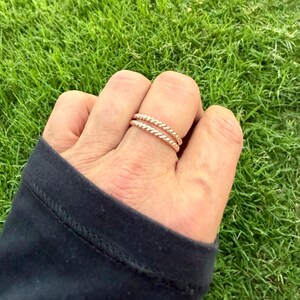 May include: Two rose gold rings with a twisted rope design are worn on a finger. The rings are set against a background of green grass.