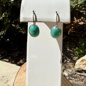 May include: A pair of turquoise earrings with a silver-colored metal setting. The earrings have a round, polished turquoise stone and a simple hook closure. The earrings are displayed on a white stand.