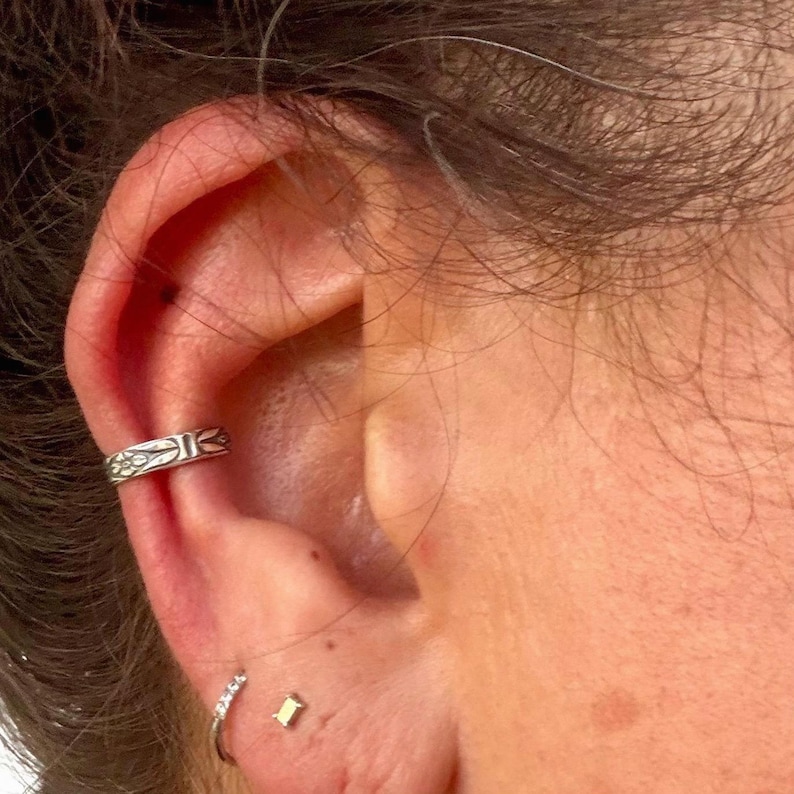 K&ouml;nnte beinhalten: Ein silberfarbener Ohrring-Cuff mit eingraviertem Blumenmuster, der am Helix getragen wird. Unterhalb des Cuffs sind ein kleiner silberner Creolen-Ohrring mit klaren Steinen sowie ein winziger goldfarbener Ohrstecker am Ohrl&auml;ppchen zu sehen.