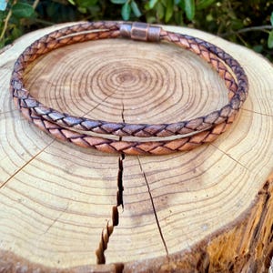 Puede incluir: Un collar de cuero trenzado de doble hebra en un degradado de tonos marr&oacute;n claro y oscuro. El collar cuenta con un cierre met&aacute;lico cil&iacute;ndrico con acabado en tono cobre que une los dos cordones de cuero trenzado en un bucle circular.