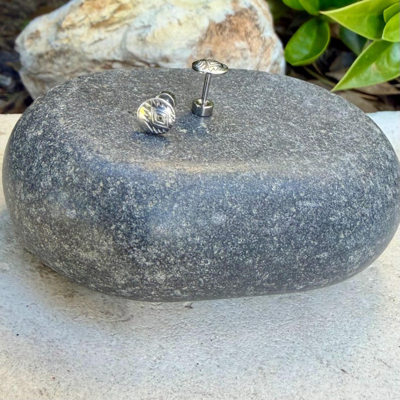 May include: A pair of silver-toned stud earrings featuring a circular face with a central diamond-shaped engraving and a thin post backing. The earrings have a metallic finish and a small, flat, round design that sits flush against the surface.