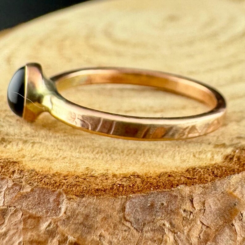 May include: A gold-coloured ring featuring a black, oval gemstone. The band has a textured pattern. The ring is displayed on a wooden surface.