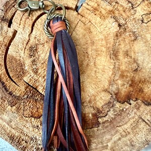 May include: A leather tassel keychain with a bronze-colored clasp and ring. The tassel features strips of dark brown, black, and tan leather. The keychain is set against a textured, wooden background.