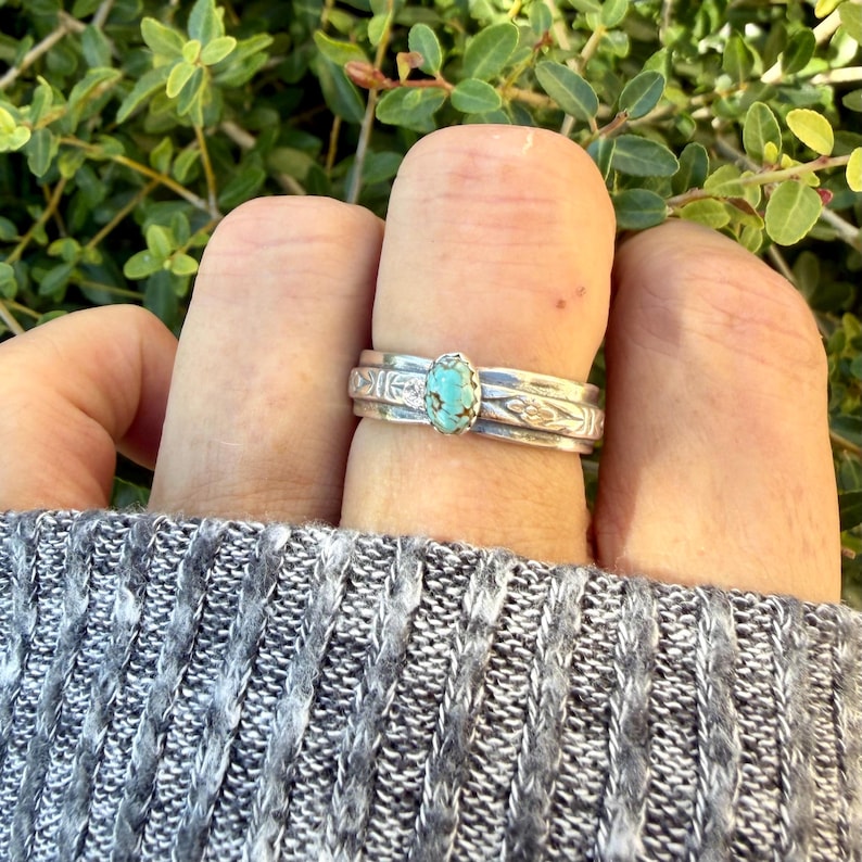 Puede incluir: Un anillo de plata con una piedra turquesa ovalada central con patrones de matriz oscuros. La banda se compone de tres segmentos de plata apilados, con las bandas exteriores mostrando patrones florales grabados en la superficie del metal.