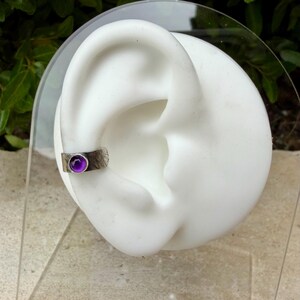May include: A silver ear cuff with a textured finish and a round, purple gemstone. The cuff is shown on a white ear model against a blurred green background. The gemstone is approximately 0.25 inches in diameter.