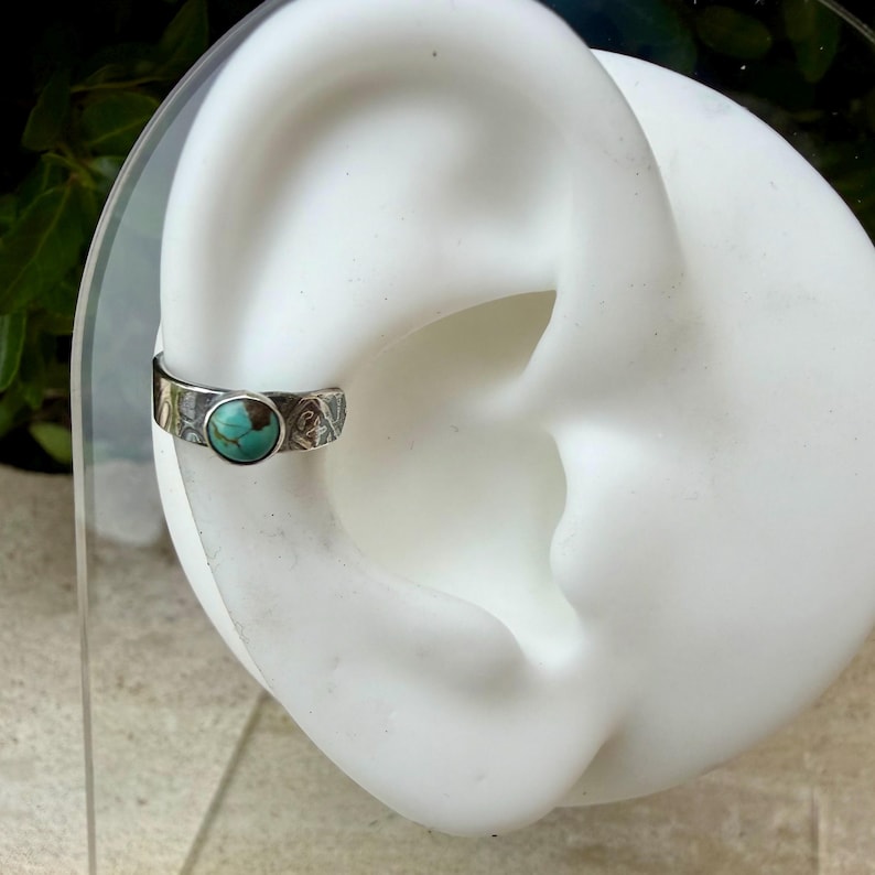 K&ouml;nnte beinhalten: Ein silberner Ohrklemmen-Schmuck mit einem T&uuml;rkisstein. Der Schmuck wird auf einem wei&szlig;en Ohrmodell pr&auml;sentiert. Das Metall hat ein strukturiertes Muster mit ge&auml;tzten Details. Der T&uuml;rkisstein ist rund und mittig platziert.