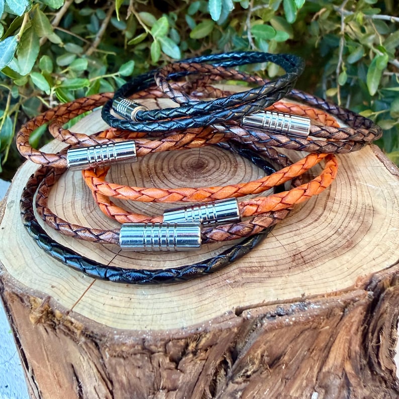 May include: A collection of braided leather bracelets in black, tan, and brown tones. Each bracelet features a cylindrical silver-toned metal magnetic clasp with a ribbed texture. The bracelets are arranged in a stacked, overlapping circular formation.