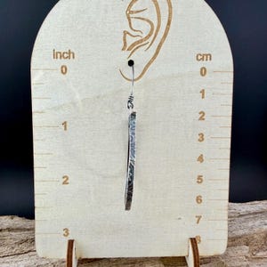 May include: A single silver earring with a textured, rectangular design. The earring is approximately 2 inches long. The earring hangs from a small hook. The earring is displayed against a wooden measuring board.