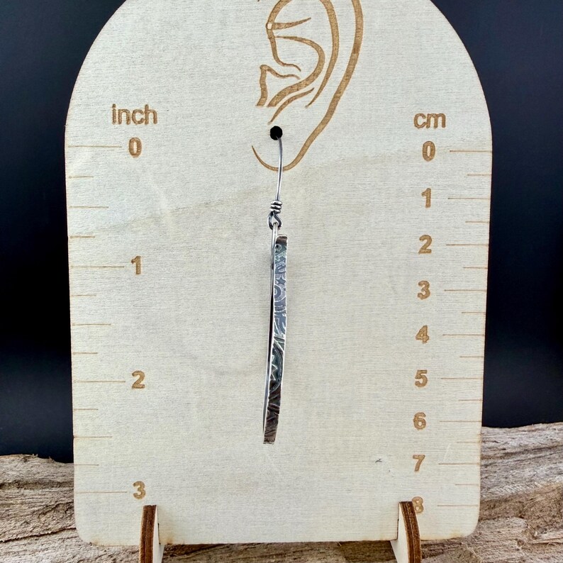May include: A single silver earring with a textured, rectangular design. The earring is approximately 2 inches long. The earring hangs from a small hook. The earring is displayed against a wooden measuring board.