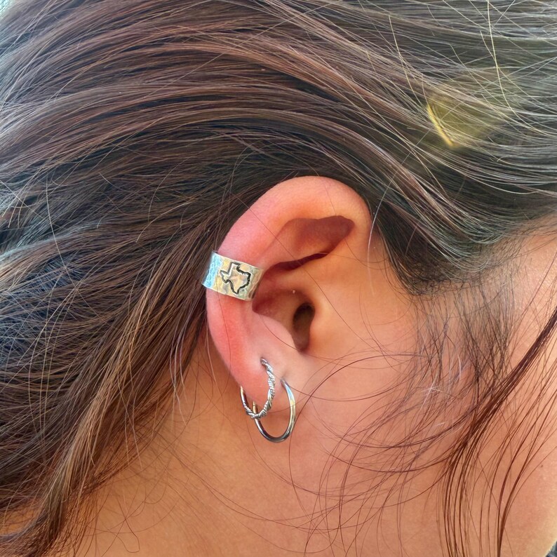 May include: A silver ear cuff featuring a stamped outline of the state of Texas on a textured surface, worn alongside two small silver hoop earrings. The ear cuff has a wide band design, while the hoops consist of one smooth and one twisted wire.