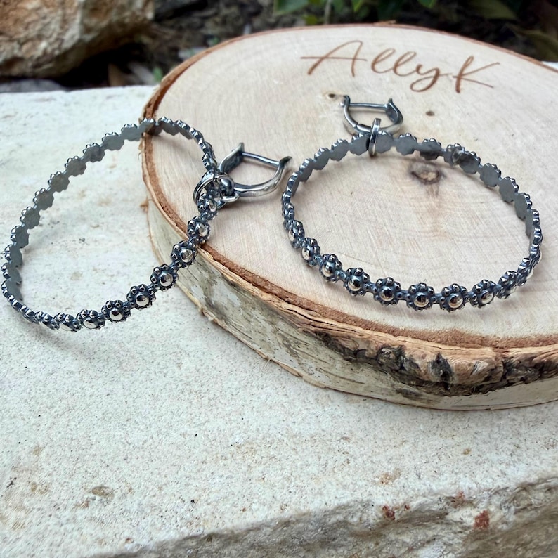 May include: A pair of silver hoop earrings with a floral design. The earrings are displayed on a wooden surface. The earrings have a dark, oxidized finish and a hinged clasp.
