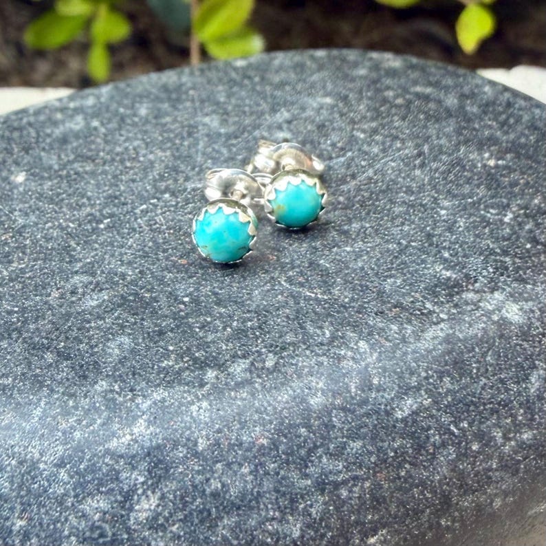 以下が含まれることがあります： ターコイズスタッドピアス。各イヤリングは、銀色のスカラップベゼルにセットされた丸い明るい青緑色の石が特徴です。イヤリングは、暗い灰色のテクスチャード加工された表面に表示されます。